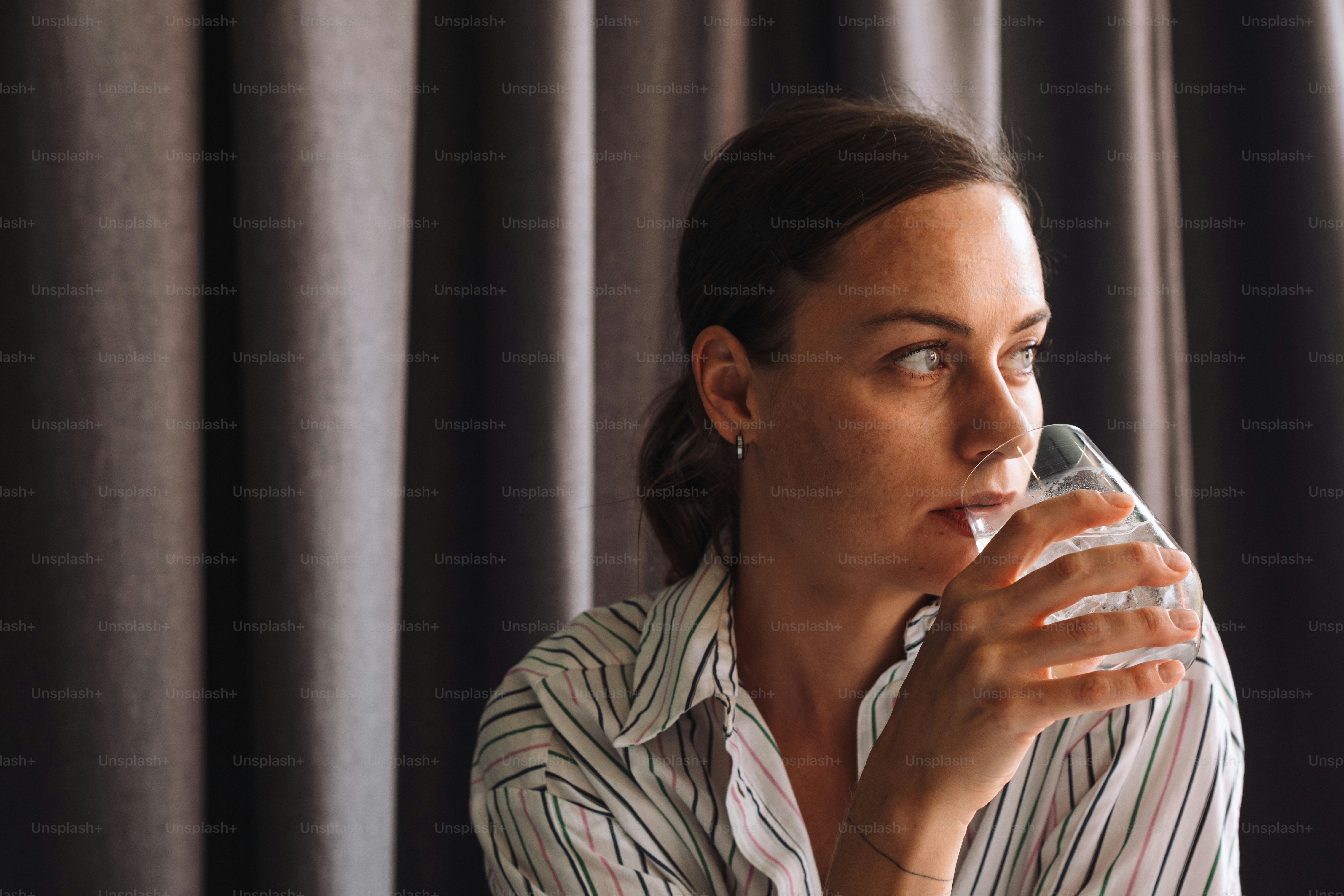 a woman drinking from a glass in front of a curtain