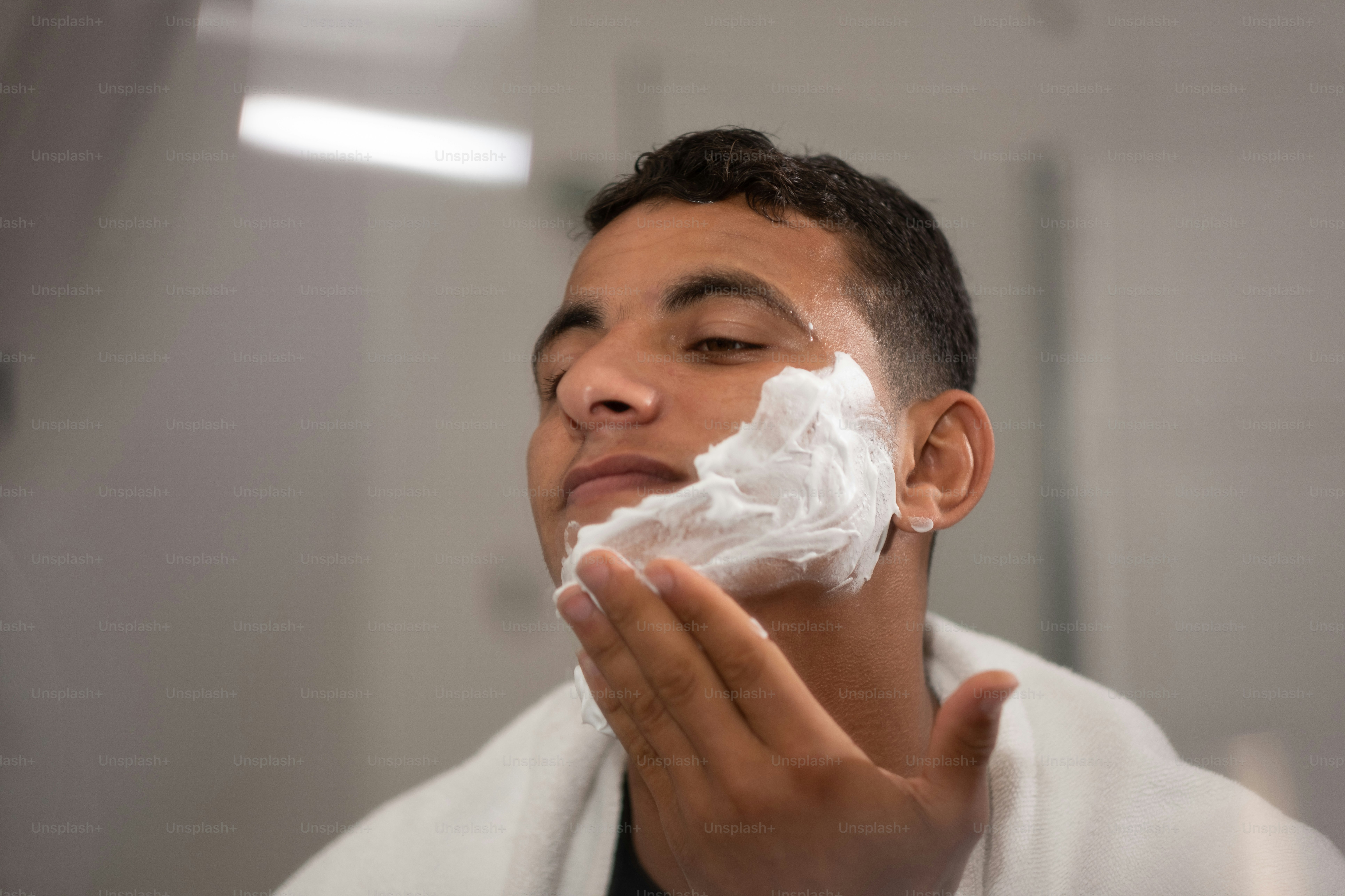 With his face covered in shaving cream, a young man looks at his reflection in the restroom mirror while shaving.