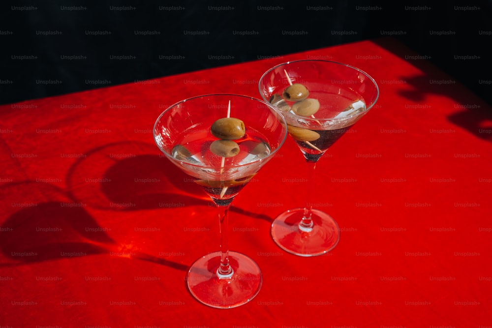 two martini glasses with olives in them on a red table
