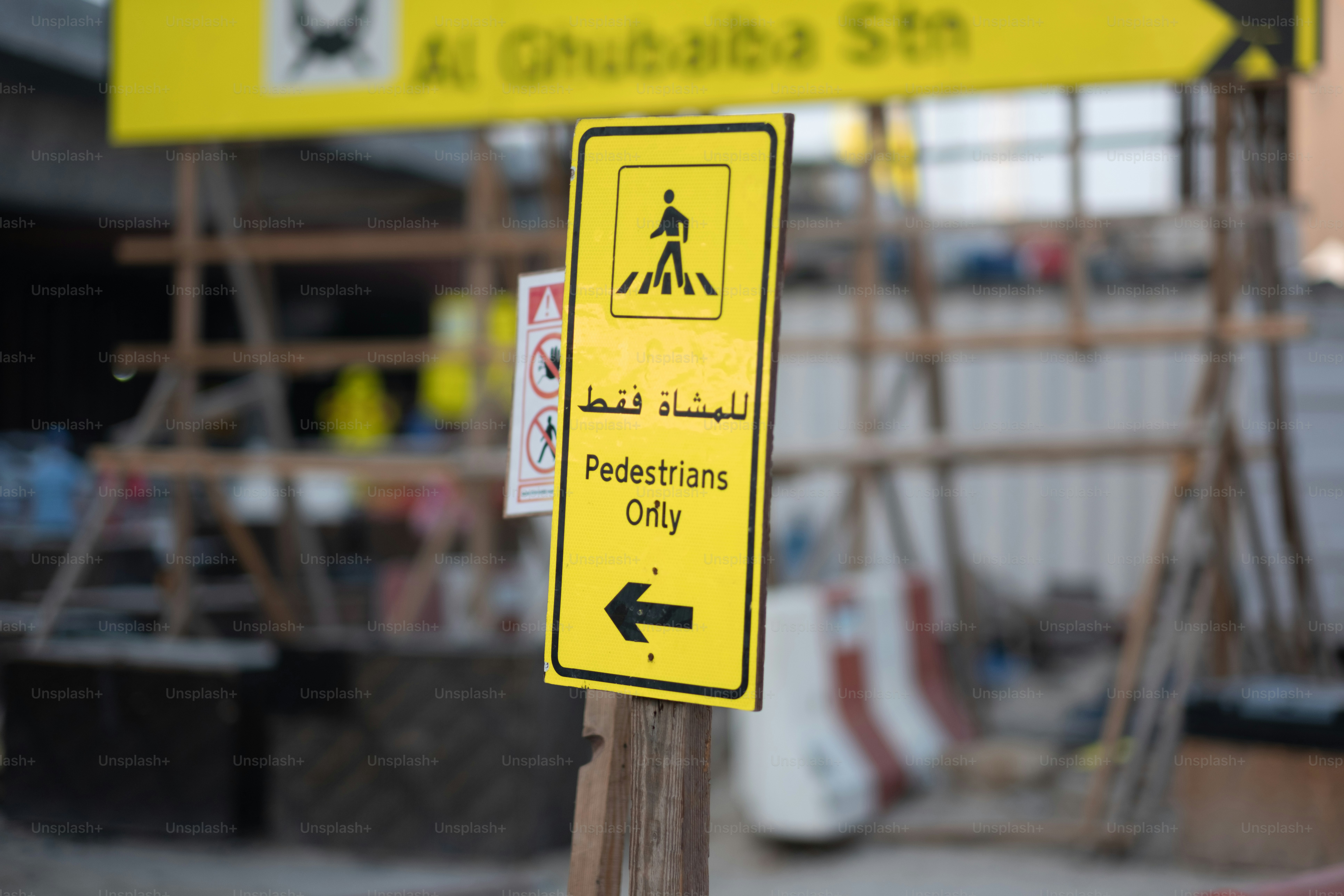 A walkway for pedestrians at a construction site, created using high-visibility red and white jersey barriers, and posted with a bilingual English and Arabic sign