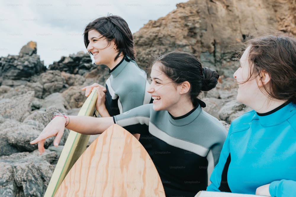 three women in wetsuits holding surfboards on a rocky beach