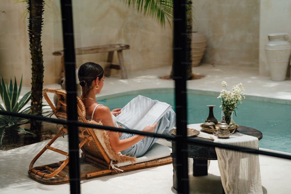 a woman sitting in a chair by a pool reading a book