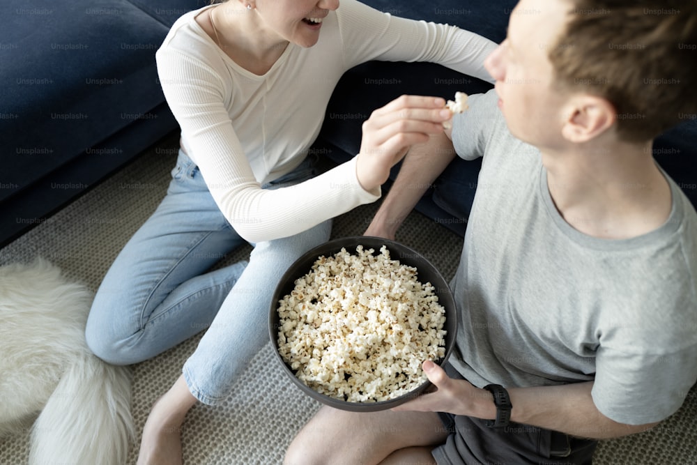 a man and woman sitting on the floor eating popcorn