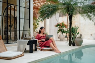 a woman sitting on a chair next to a pool using a laptop