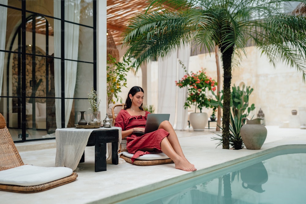 a woman sitting on a chair next to a pool using a laptop