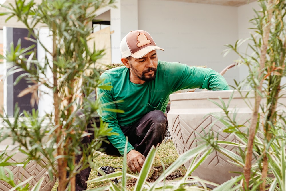 a man in a green shirt and a hat crouching next to a plant