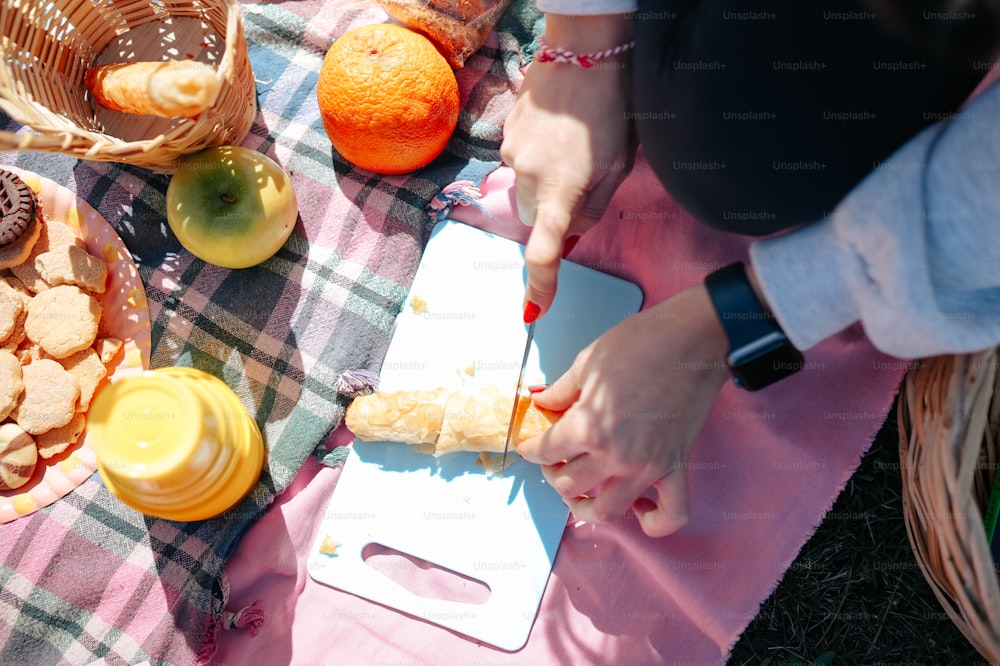 a woman is cutting a sandwich on a picnic blanket