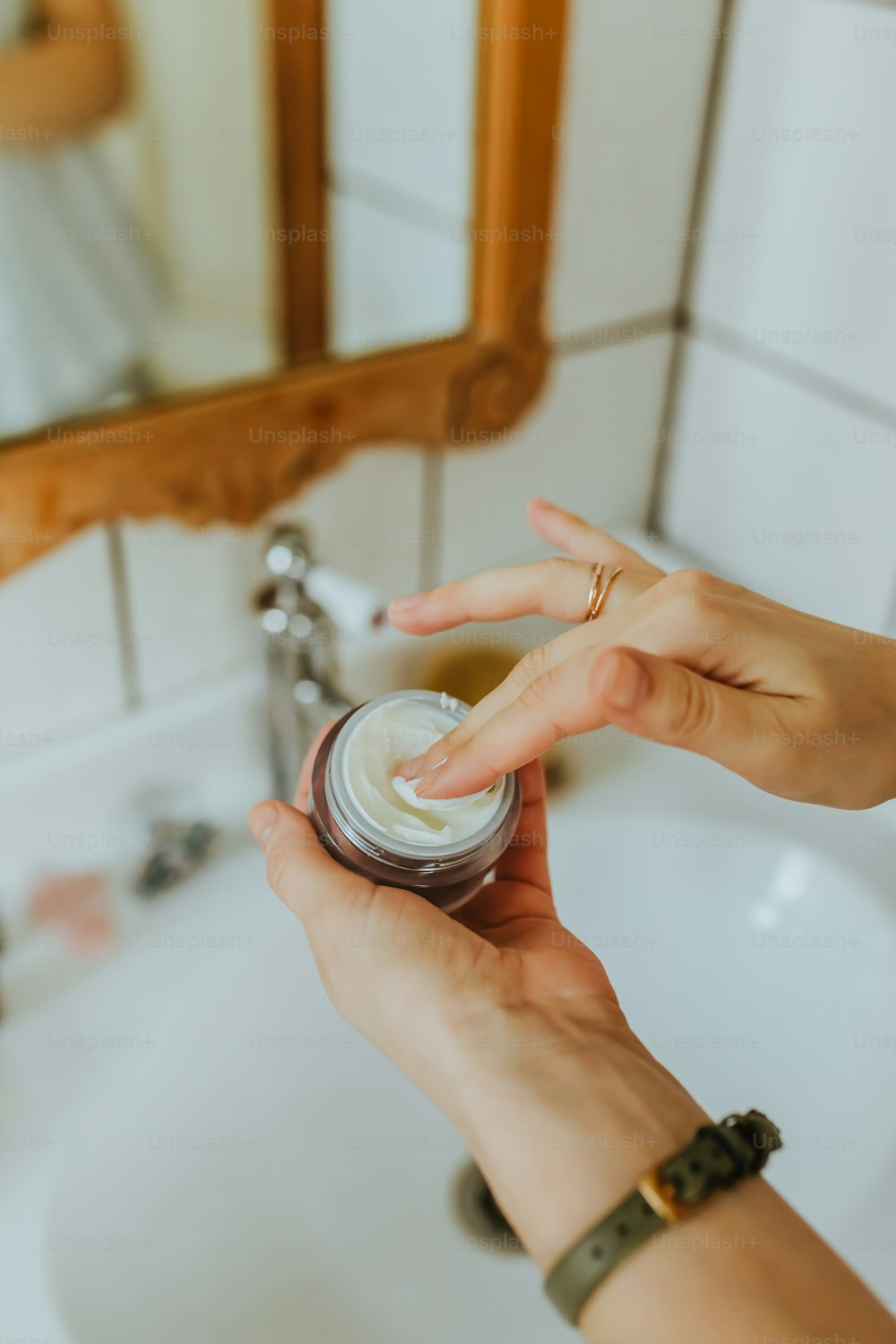 a person holding a jar of cream in front of a bathroom sink