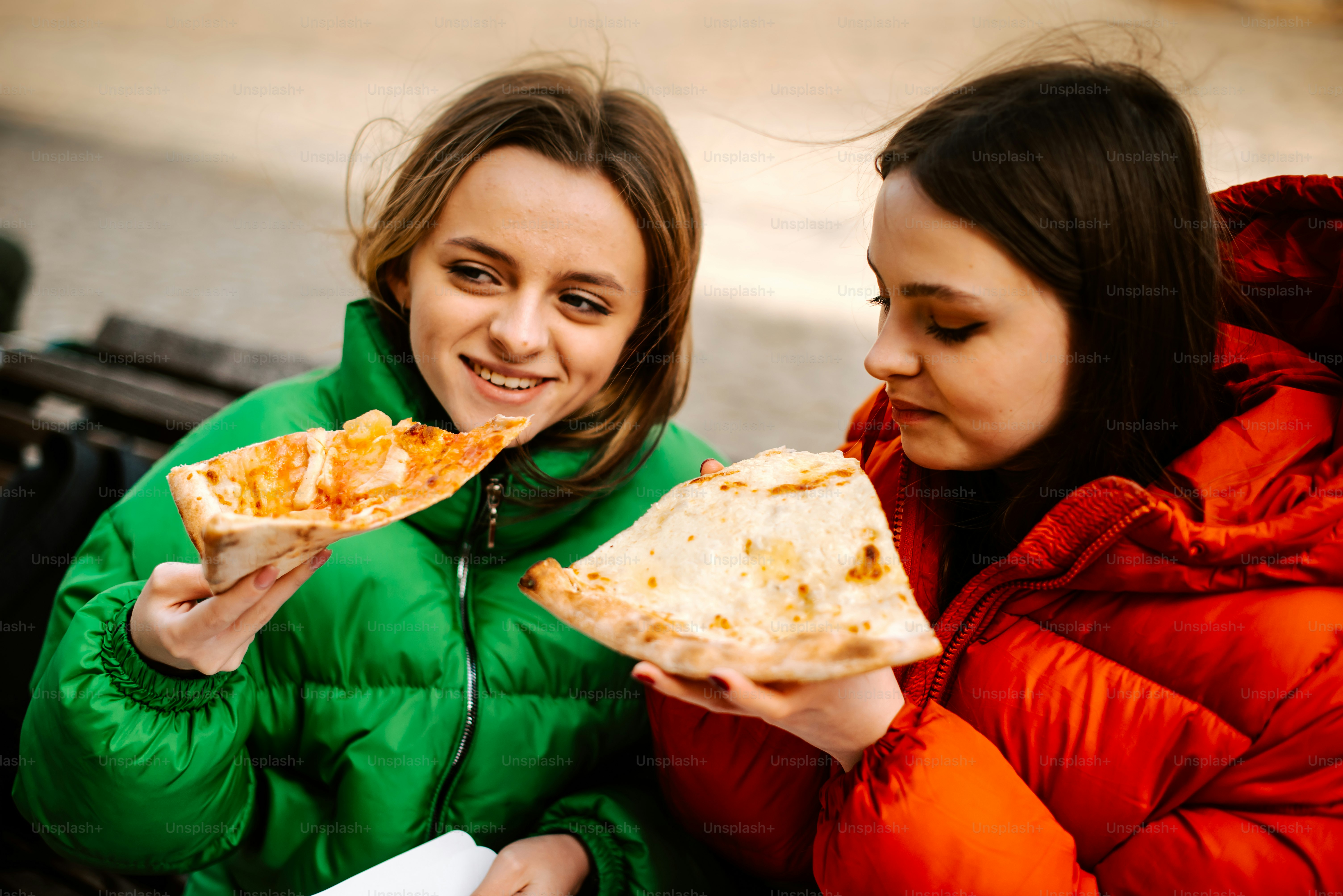 two women sitting on a bench eating pizza