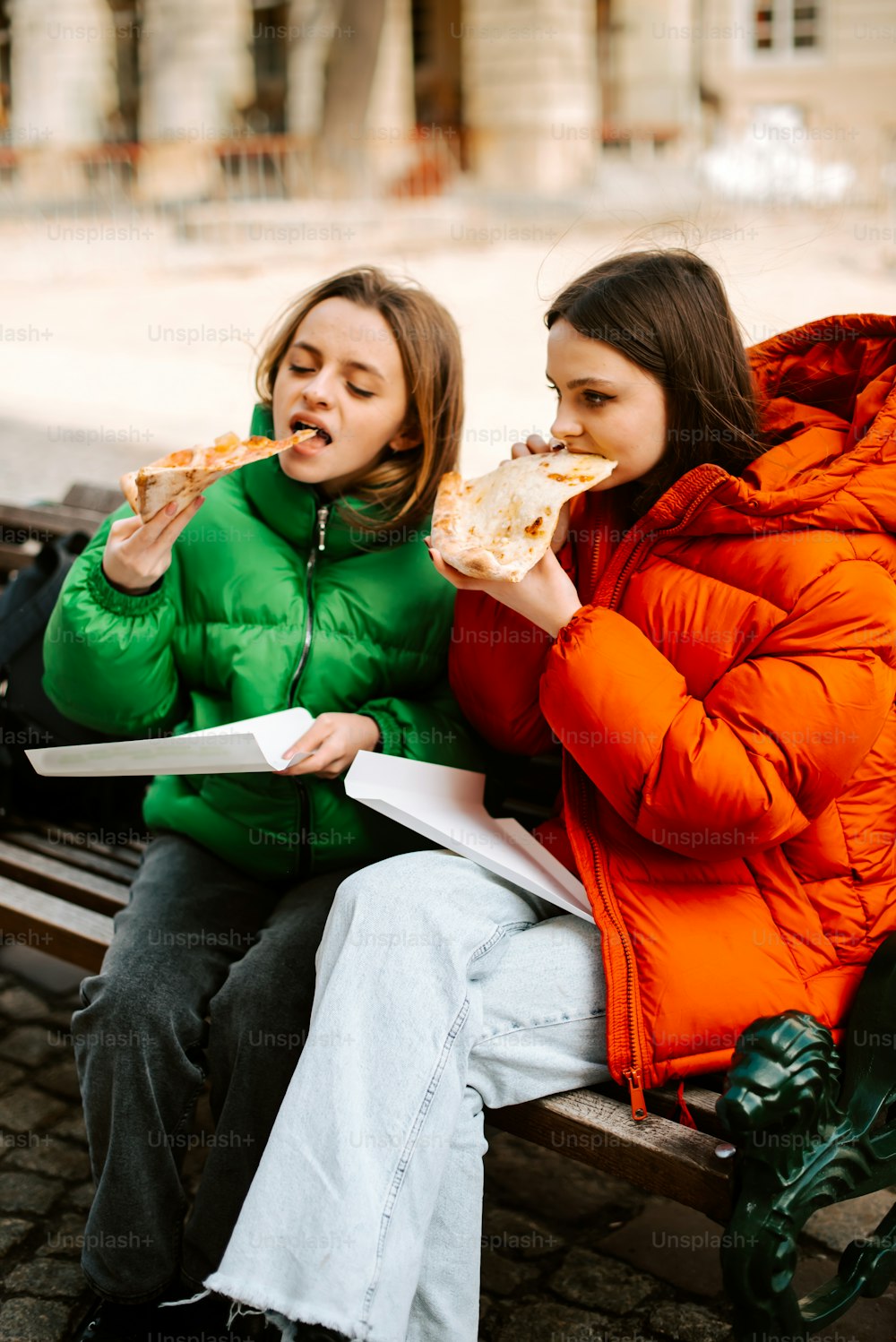 two women sitting on a bench eating pizza