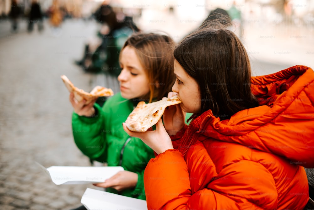 two women sitting on a bench eating pizza