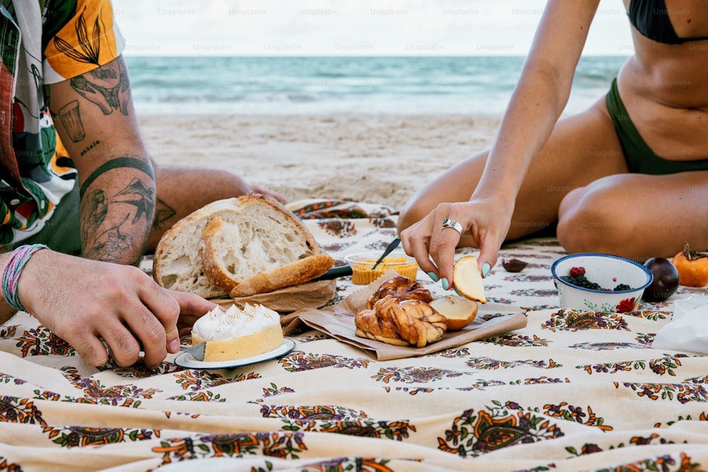 a man and woman sitting on the beach eating food