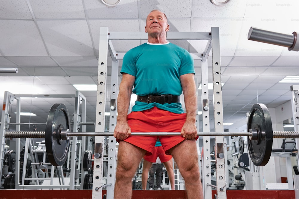 Un hombre con camisa verde y pantalones cortos rojos levanta una barra en un gimnasio