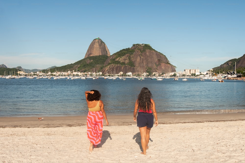 two women walking on a beach next to a body of water