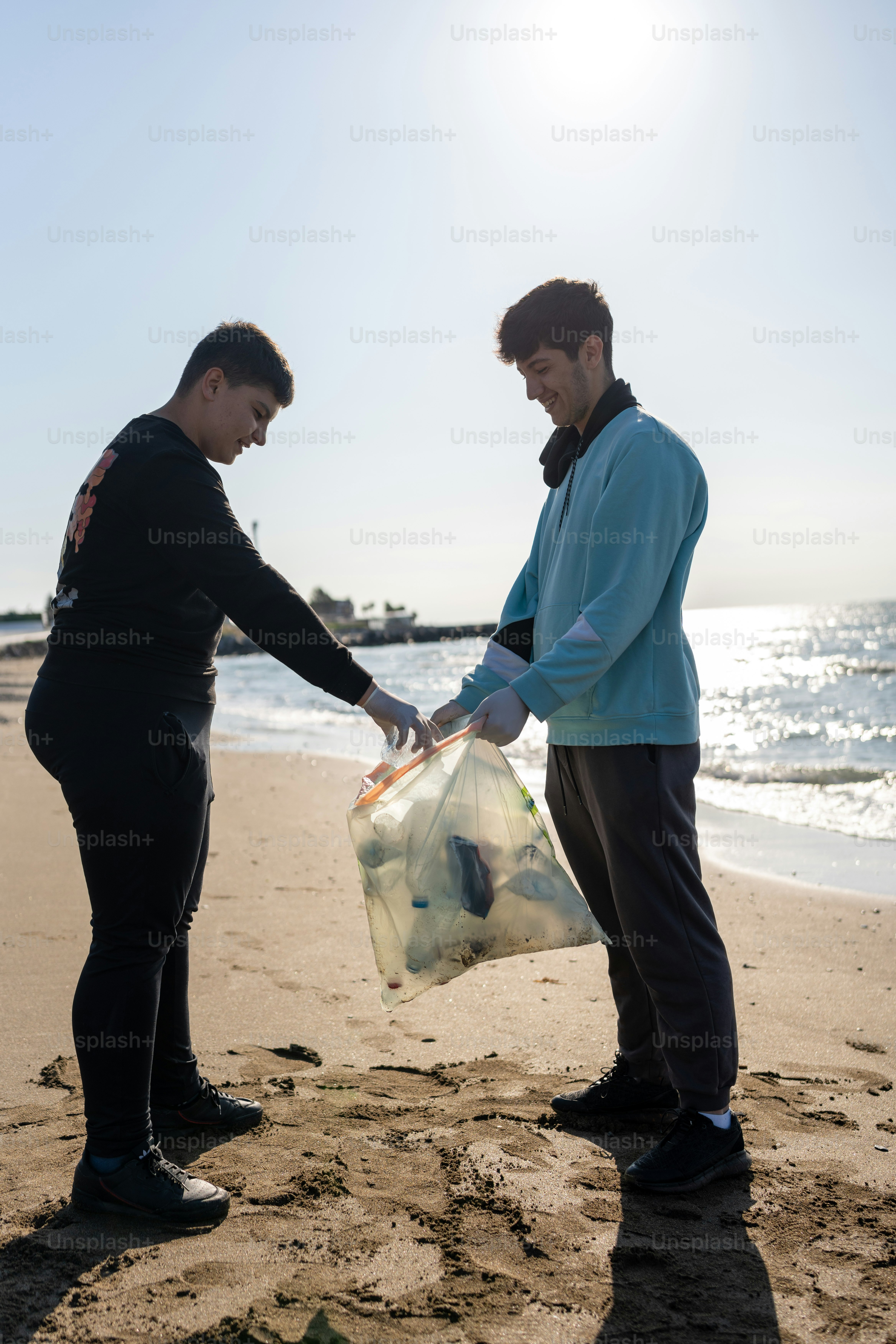 two men standing on a beach holding a plastic bag