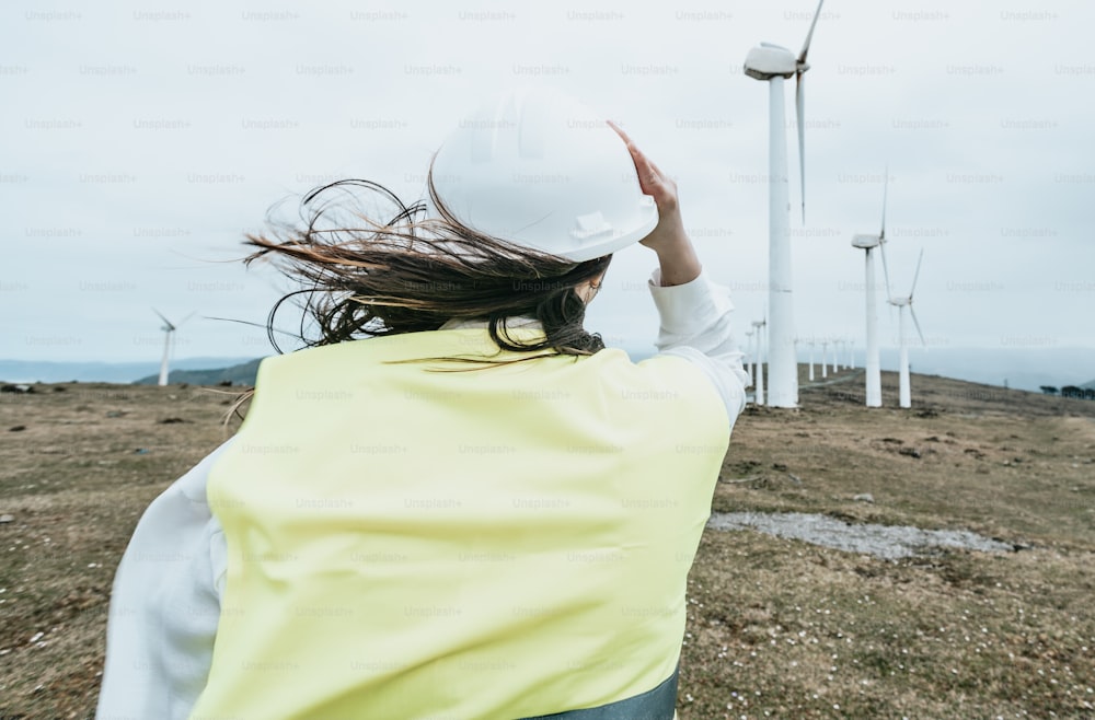 Una mujer con un chaleco amarillo y un casco blanco parada frente a las turbinas eólicas