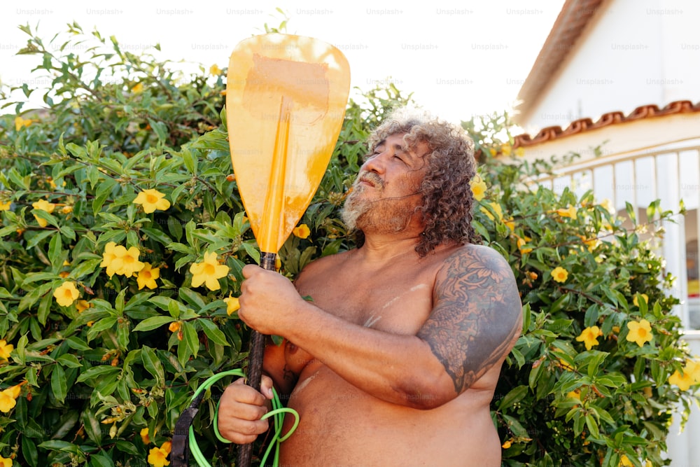 a shirtless man holding a yellow paddle in front of a bush