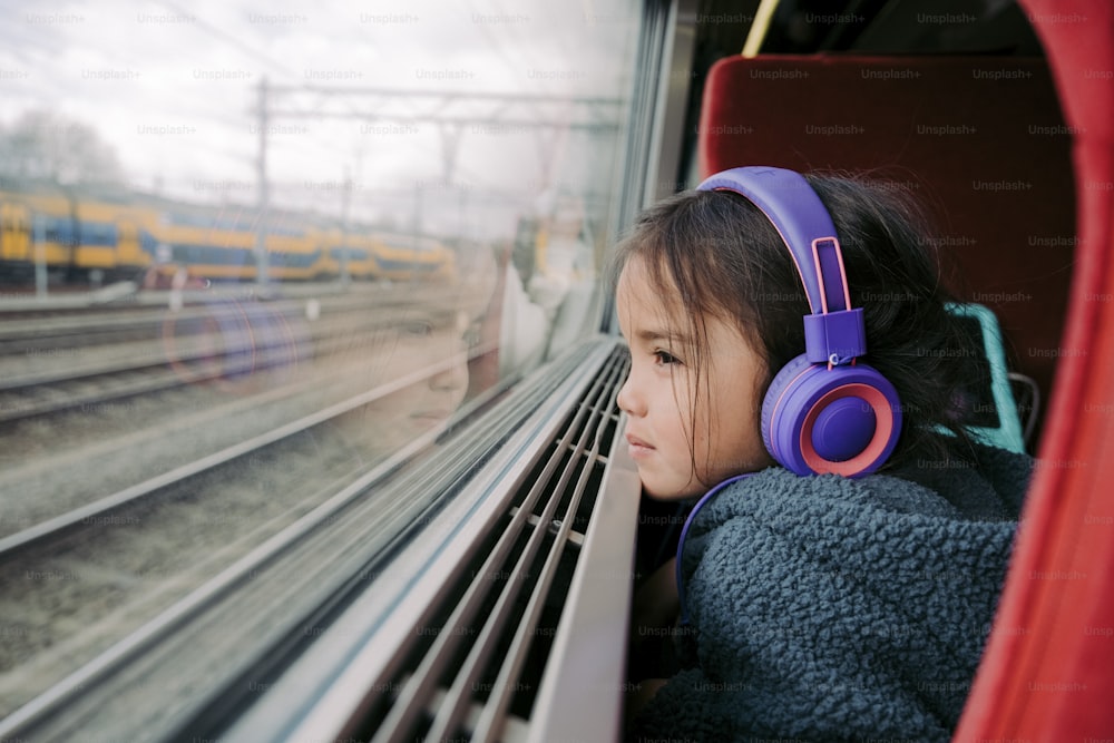 a little girl wearing headphones looking out a train window