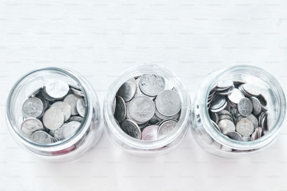 três frascos de vidro cheios de moedas em uma superfície branca