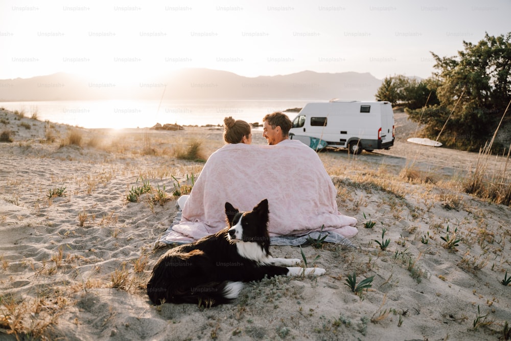 a man and woman sitting in the sand with a dog