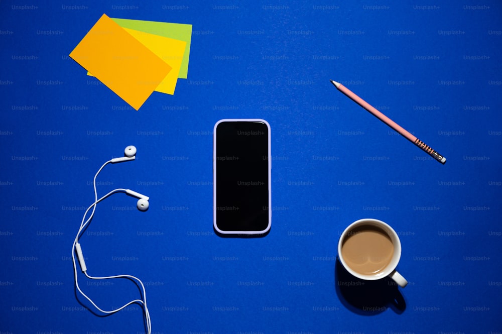 uma xícara de café, fones de ouvido e um telefone celular em uma superfície azul