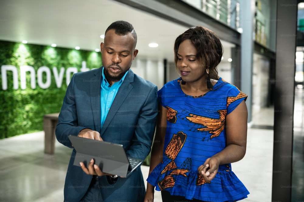 Two young African businesspeople discussing work together over a digital tablet while walking along an office hallway
