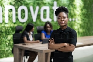 Selbstbewusste junge afrikanische Geschäftsfrau steht mit verschränkten Armen in einem Büro-Sitzungssaal, während Kollegen im Hintergrund arbeiten