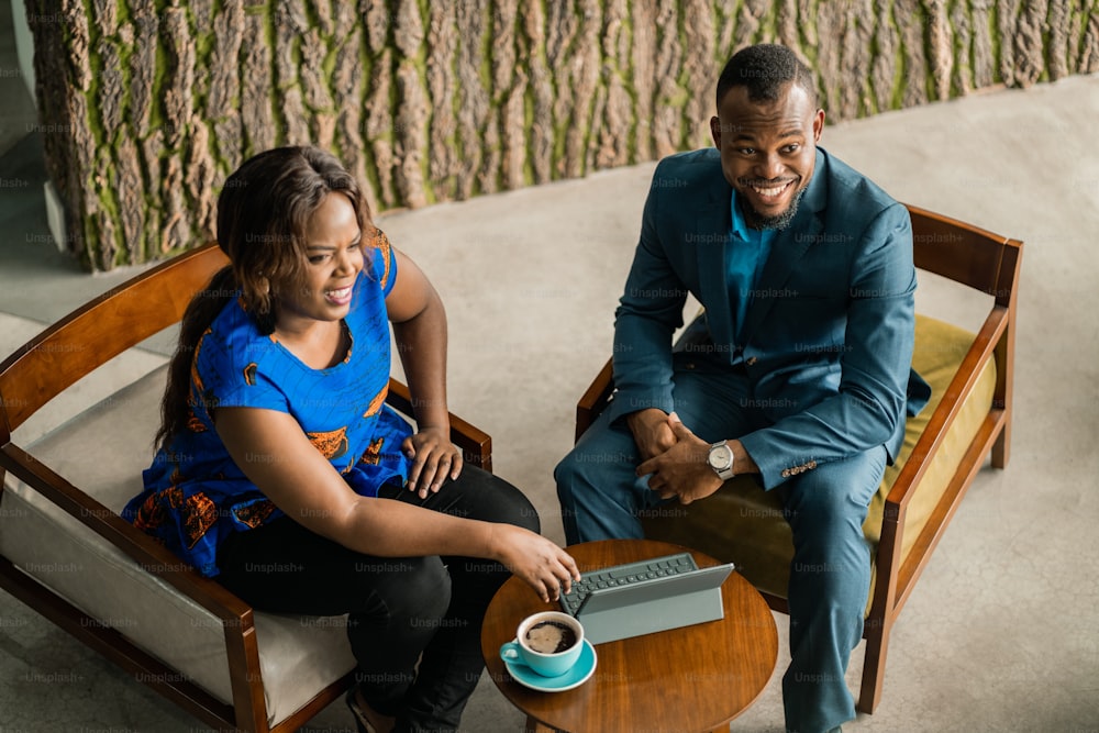 Angolo alto di due giovani imprenditori africani sorridenti che parlano insieme su una tavoletta digitale durante una riunione informale in un salotto dell'ufficio