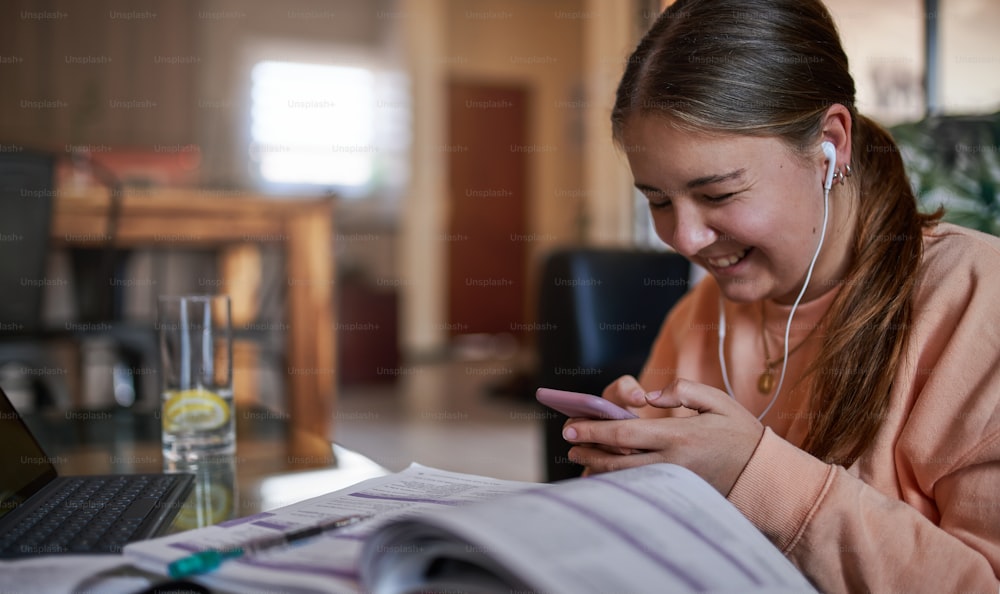 Retrato de una hermosa y sonriente joven adolescente sentada en la mesa de café mensajería, usando un teléfono inteligente en el acogedor interior de la sala de estar, estudiando y relajándose en casa