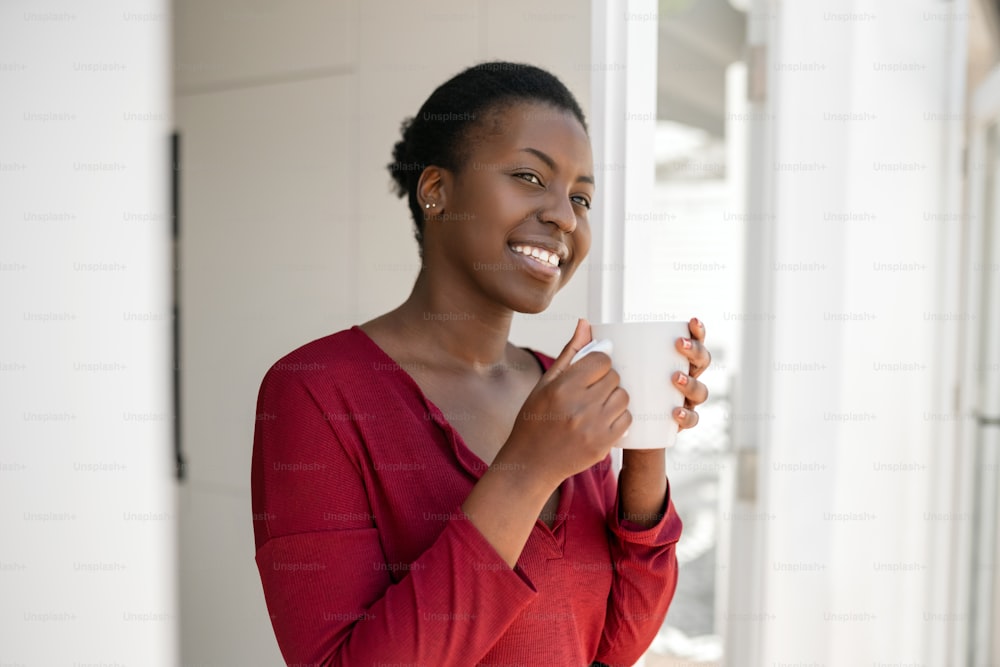 Retrato de una hermosa mujer africana parada en la puerta de su casa, sosteniendo una taza, sonriendo y mirando a lo lejos