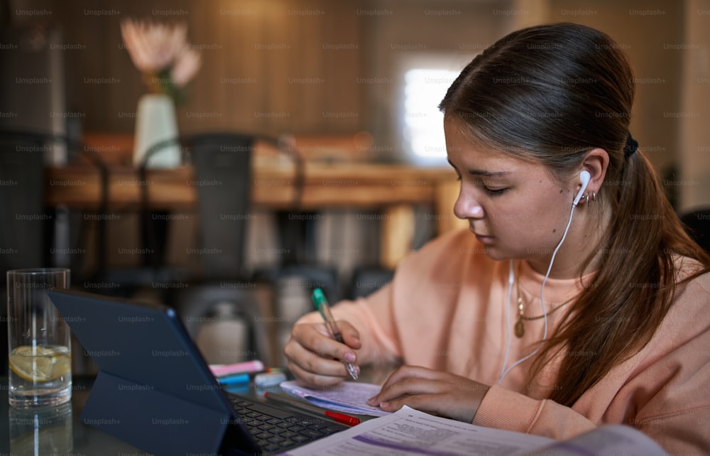 Retrato de una joven adolescente de la escuela sentada en la mesa de café escuchando música, usando auriculares en el interior moderno y acogedor de la sala de estar, estudiando y relajándose en casa. Educación en línea y concepto de aprendizaje electrónico durante la cuarentena