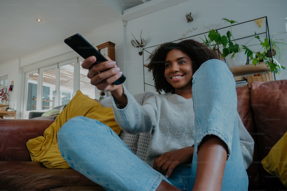 mixed-race woman holding the television remote and changing the channel on the couch while smiling . High quality photo