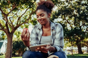 Adolescente de raza mixta investigando para un proyecto en tableta digital mientras sostiene tomate fresco agachado en campos deliciosos. Foto de alta calidad