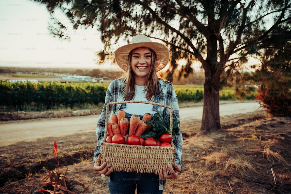 Agricultora caucásica riendo y sonriendo mientras sostiene una canasta fresca de verduras trabajando al amanecer en la granja. Foto de alta calidad