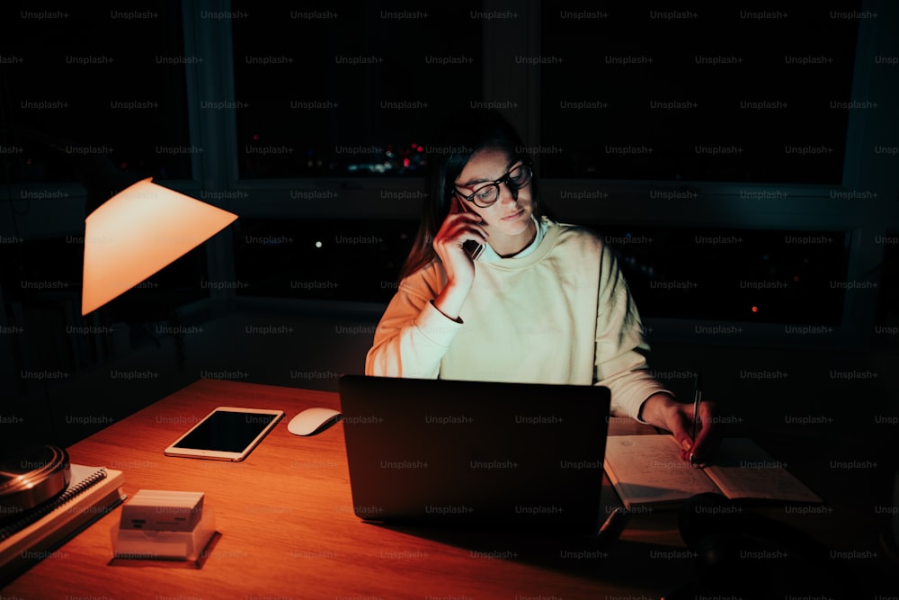 Caucasian female student chatting to colleagues on telephone working late at night sitting infront of laptop in home office. High quality photo