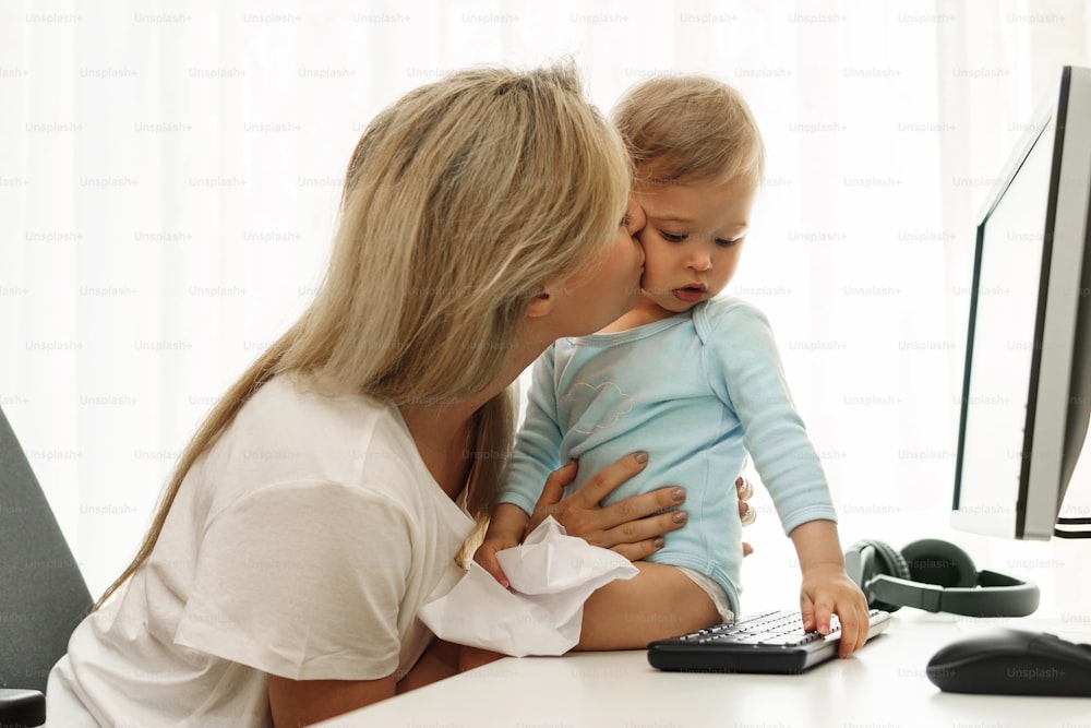 Young freelancer mother is kissing her little son sitting on the desk at her home office workplace on the cheek.