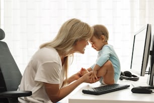 Young smiling freelancer mother is playing with her little son sitting on the desk at her home office workplace.