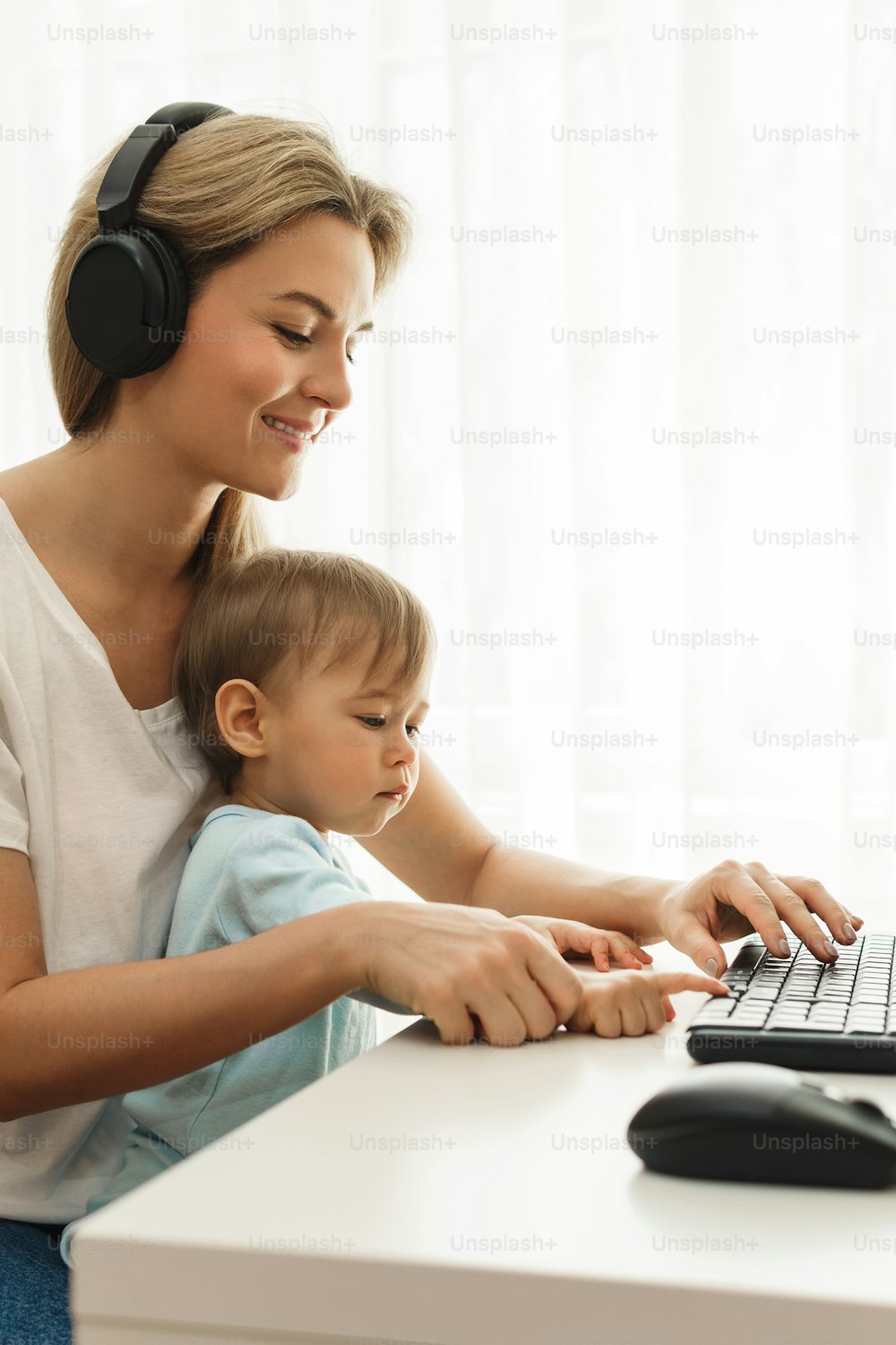 Young freelancer mother is working on computer at her home office workplace with her little son sitting on her lap.