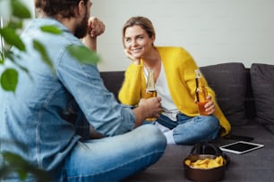 Young cheerful couple sitting on the sofa drinking beer and eating nachos at home
