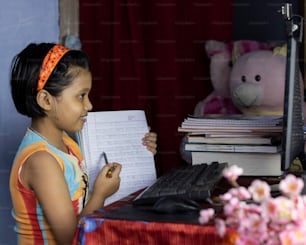 una niña india con cara sonriente asistiendo a clase en línea en una computadora de escritorio y mostrando un cuaderno a la maestra durante el brote pandémico de COVID-19