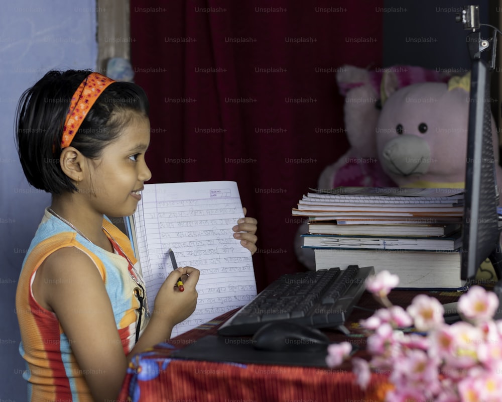 an Indian girl child with smiling face attending online class on desktop computer and showing notebook to teacher during COVID-19 pandemic outbreak