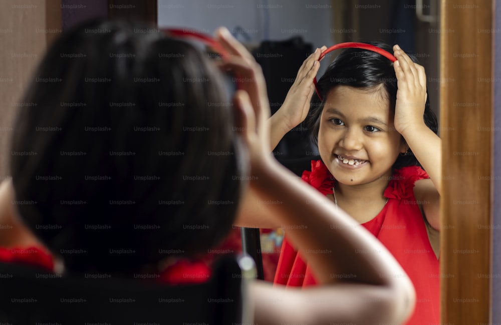 a cute Indian girl child in red dress is styling hair with hairband in front of mirror with smiling face