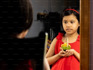 빨간 드레스를 입은 귀여운 인도 소녀가 인형을 들고 거울 앞에 서 있다