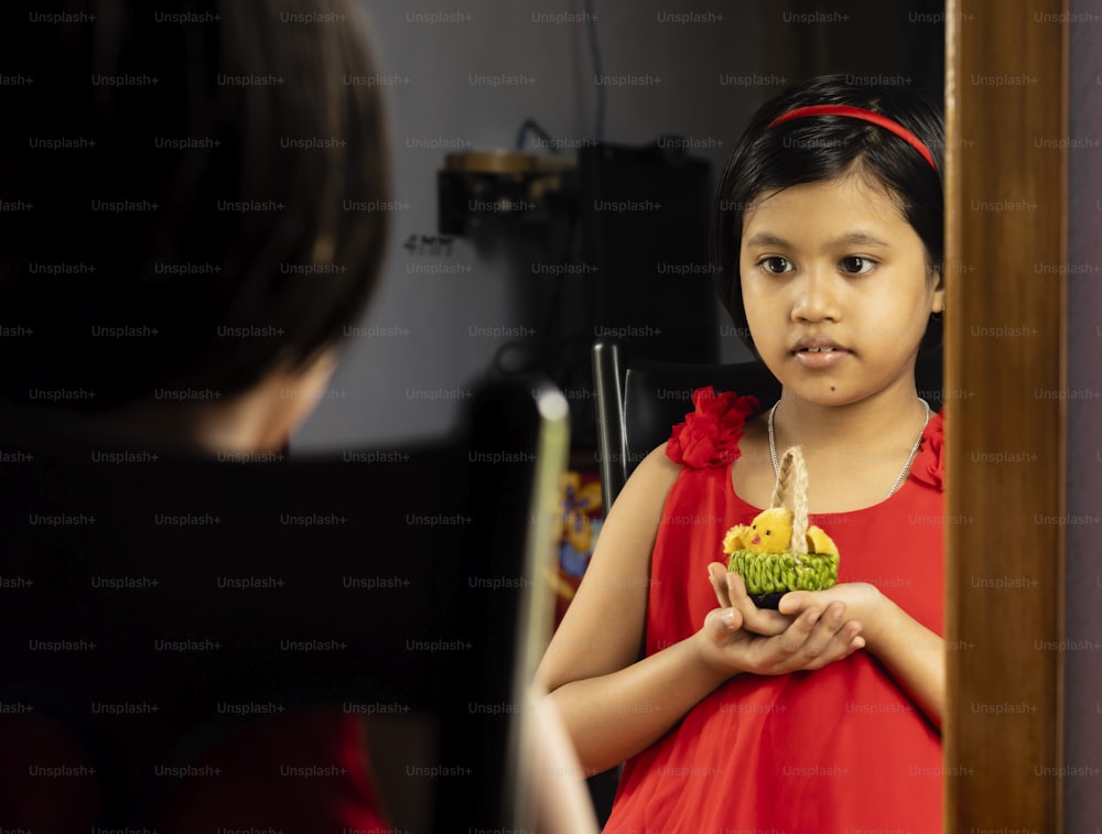 a cute Indian girl child in red dress standing in front of mirror with a doll
