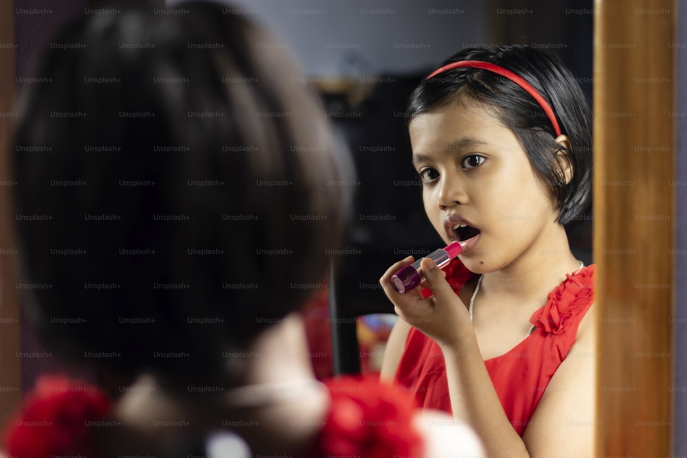 a cute Indian girl child in red dress is applying lipstick in front of mirror