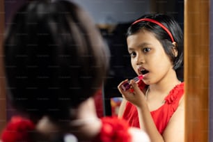 una bambina indiana carina in vestito rosso sta applicando il rossetto davanti allo specchio