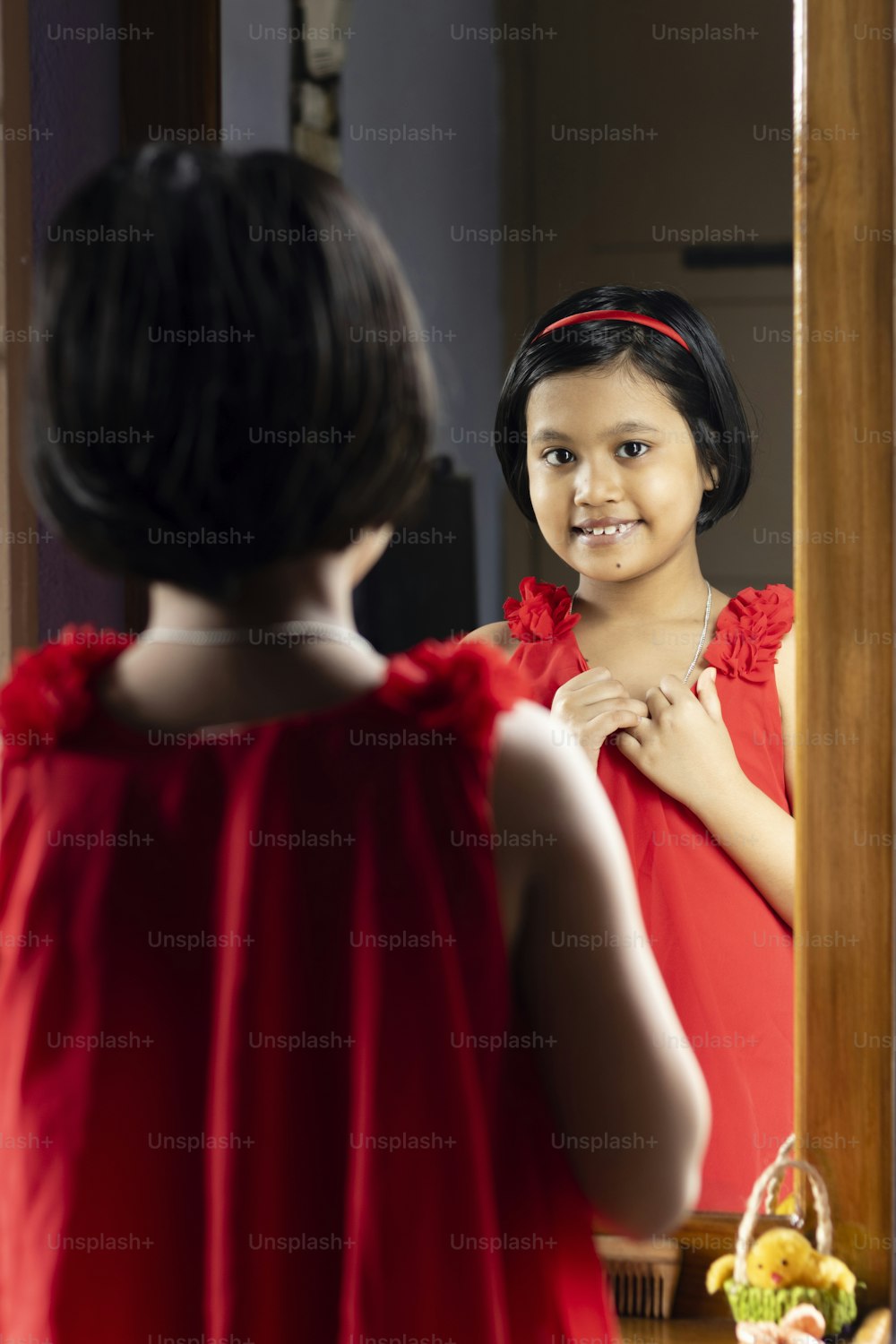 una linda niña india con vestido rojo mirando a la cámara y de pie frente al espejo con cara sonriente