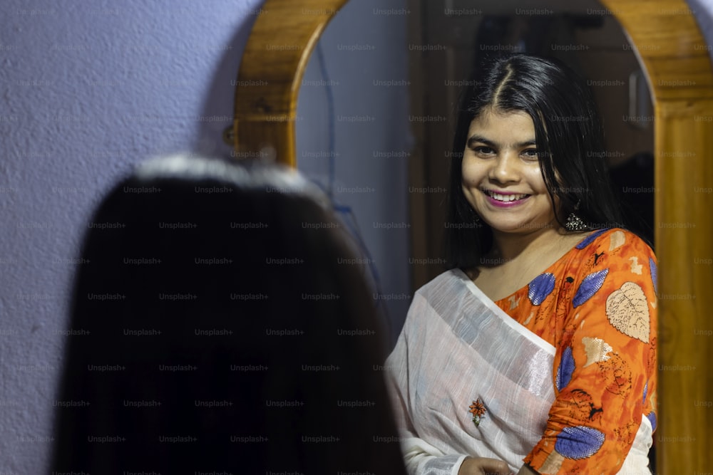 하얀 사리를 입은 아름다운 인도 여성이 웃는 얼굴로 거울 앞에서 포즈를 취하고 있다