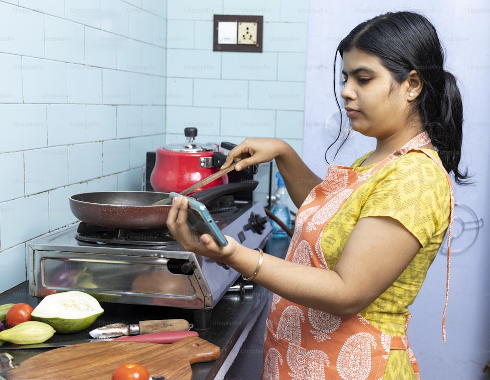 エプロンを着た美しいインドの若い女性が、ガスストーブの家庭の台所でスマートフォンで料理ビデオを見ている