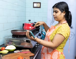 앞치마를 두른 예쁜 인도 젊은 여성이 가스레인지에 있는 가정용 주방에서 스마트폰으로 요리 비디오를 보고 있다